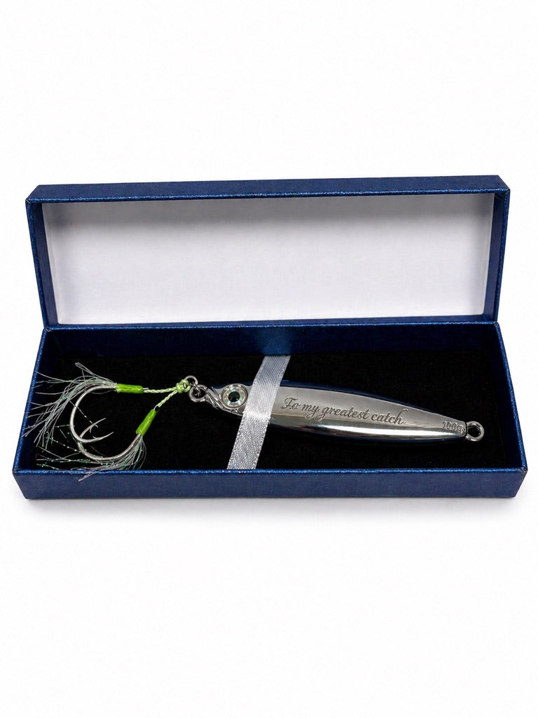 A silver metal fishing lure with a green and white skirt and treble hooks, presented inside an open dark blue gift box with a white interior and a silver ribbon.