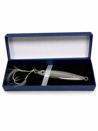 A silver metal fishing lure with a green and white skirt and treble hooks, presented inside an open dark blue gift box with a white interior and a silver ribbon.