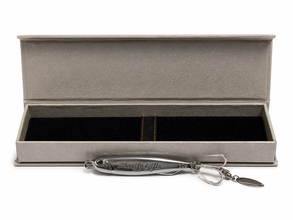 A silver metal fishing lure with a keychain attachment displayed in front of an open textured gray presentation box on a white background.