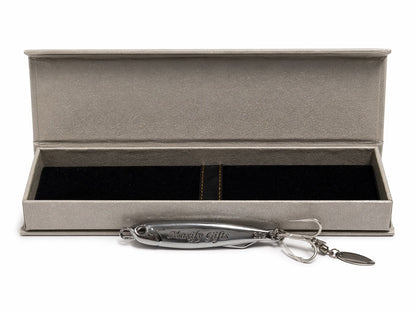 A silver metal fishing lure with a keychain attachment displayed in front of an open textured gray presentation box on a white background.