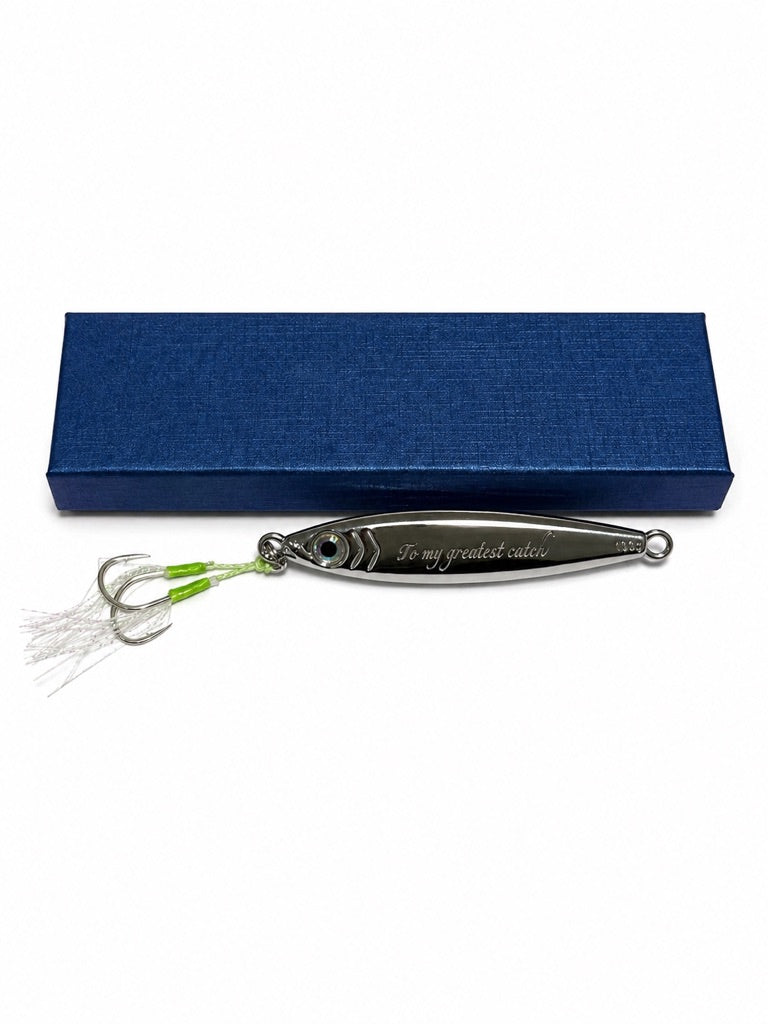 A silver metal fishing lure engraved with 'To my greatest catch' lies in front of a textured dark blue presentation box on a plain white background.