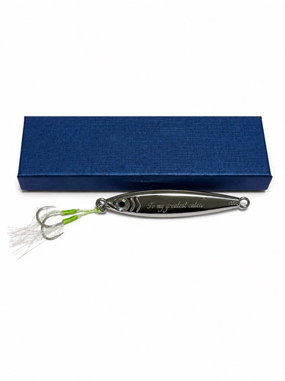 A silver metal fishing lure engraved with 'To my greatest catch' lies in front of a textured dark blue presentation box on a plain white background.