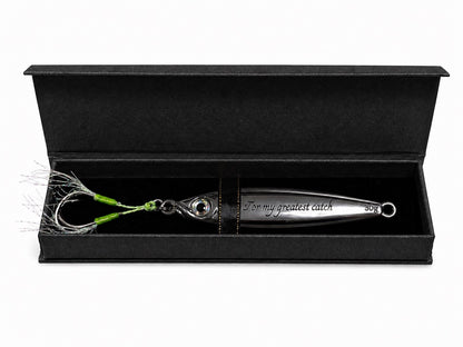 A silver metal fishing lure with green and white skirt strands, presented inside an open black gift box on a white background. The lure is engraved with the text 'For my greatest catch' and '30g'.