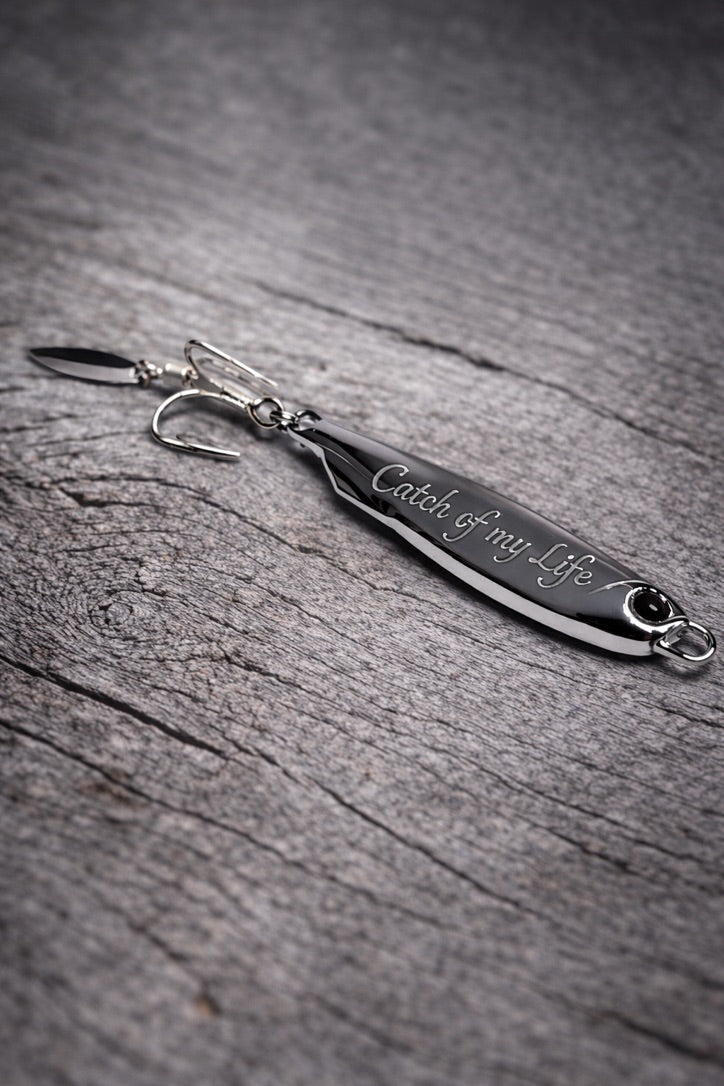 A silver metal spinnerbait fishing lure engraved with the phrase 'Catch of my life' resting on a textured gray wooden surface.