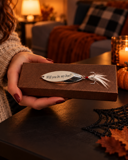 A person holds a brown gift box featuring a novelty fishing lure engraved with the phrase 'Will you be my boat?'. The scene is set in a cozy, warmly lit living room with autumn decorations, including a spiderweb, pumpkins, and a plaid throw blanket in the background.