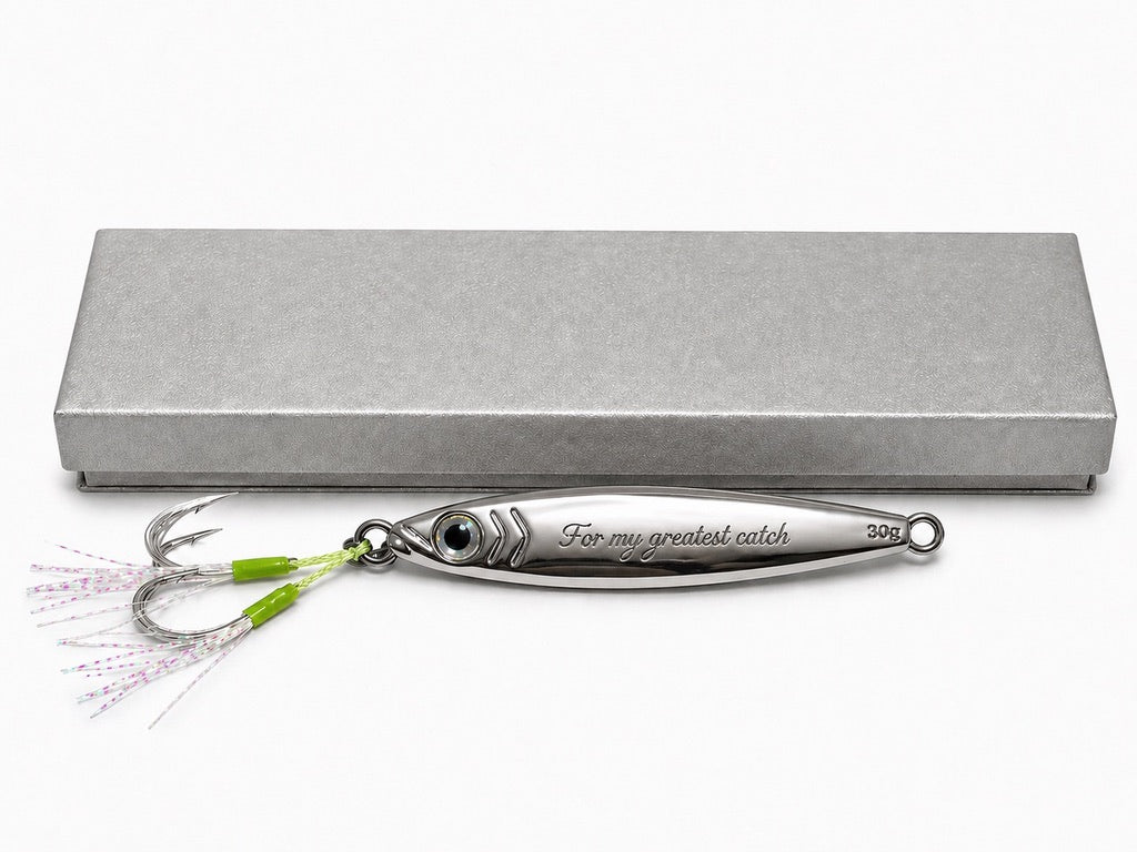 A silver metal fishing lure with a green and pink skirt and treble hooks, displayed in front of a textured gray presentation box on a white background. The lure is engraved with 'For my greatest catch'.