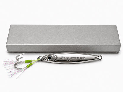 A silver metal fishing lure with a green and pink skirt and treble hooks, displayed in front of a textured gray presentation box on a white background. The lure is engraved with 'For my greatest catch'.