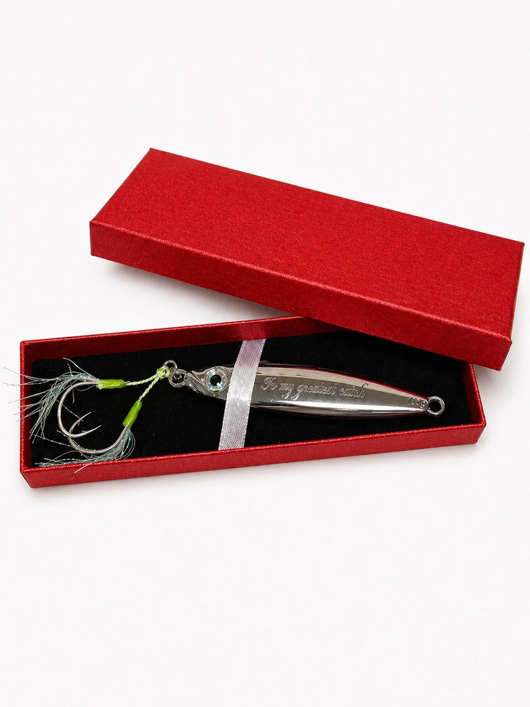 A silver metal fishing lure with a green and white skirt and a small blue eye, presented in an open red gift box with a black interior on a white background.