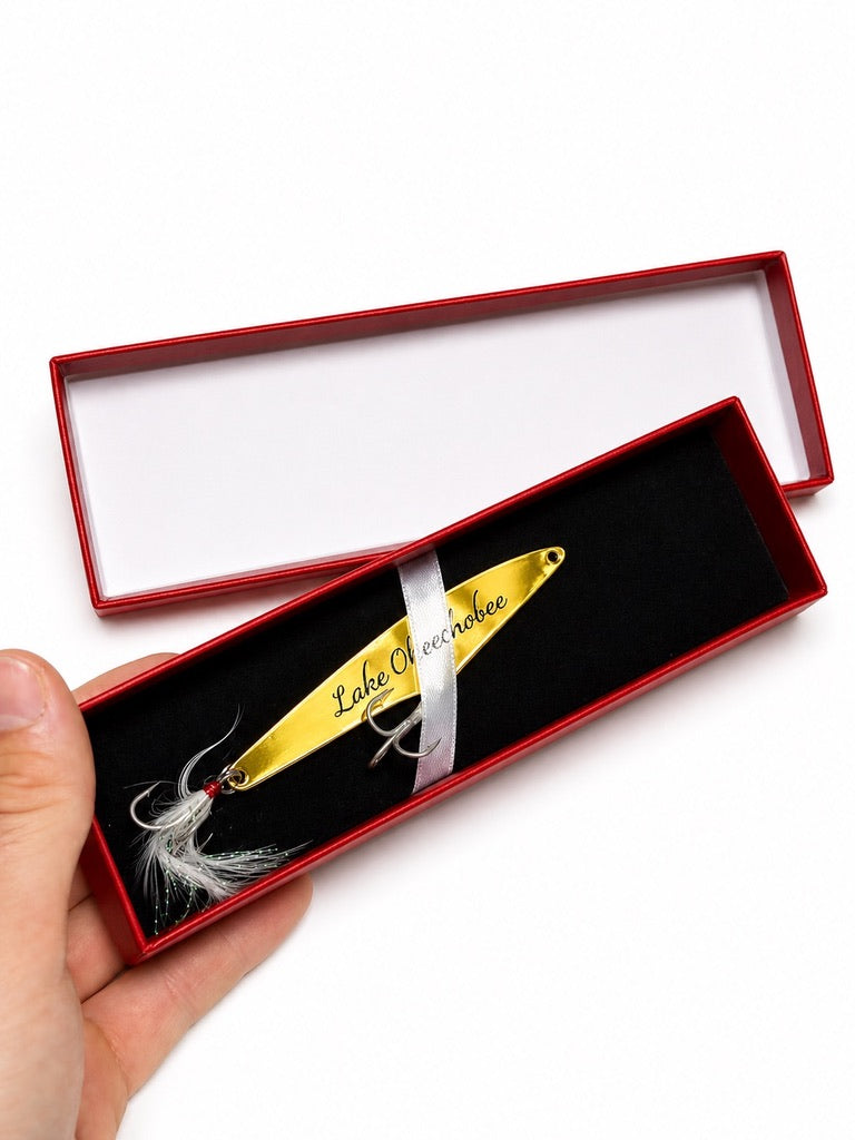 A hand holds an open red presentation box containing a gold fishing lure with 'Lake Chocó National' printed on it, alongside a white fly and a white ribbon, against a white background.