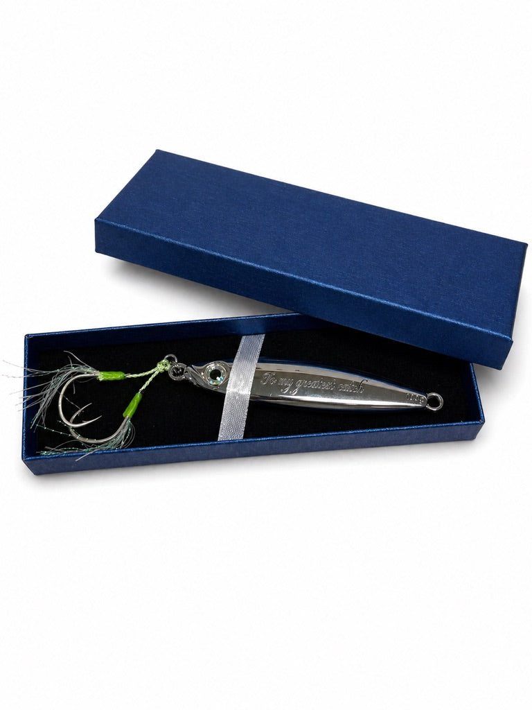 A silver metal fishing lure with green and white feathered skirt details, presented inside an open dark blue velvet gift box with its lid propped behind it on a white background.