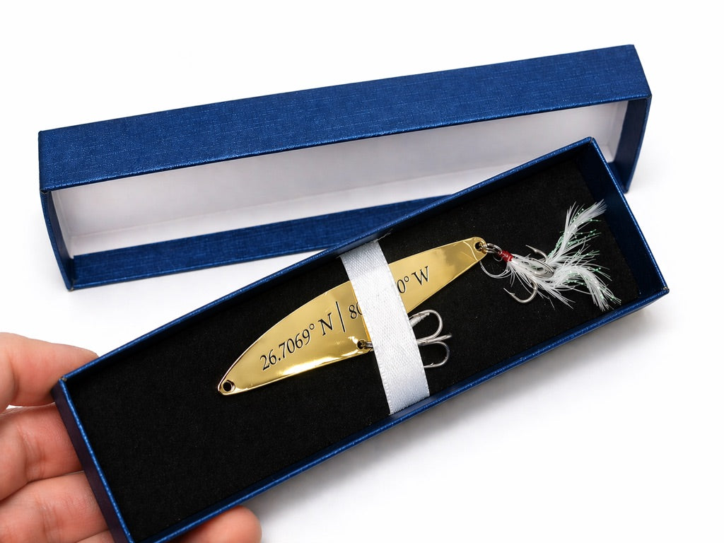 A gold metal fishing lure engraved with geographic coordinates, presented in an open blue gift box with a black insert. A hand is holding the box on the left, and the lure has white and red feather details.