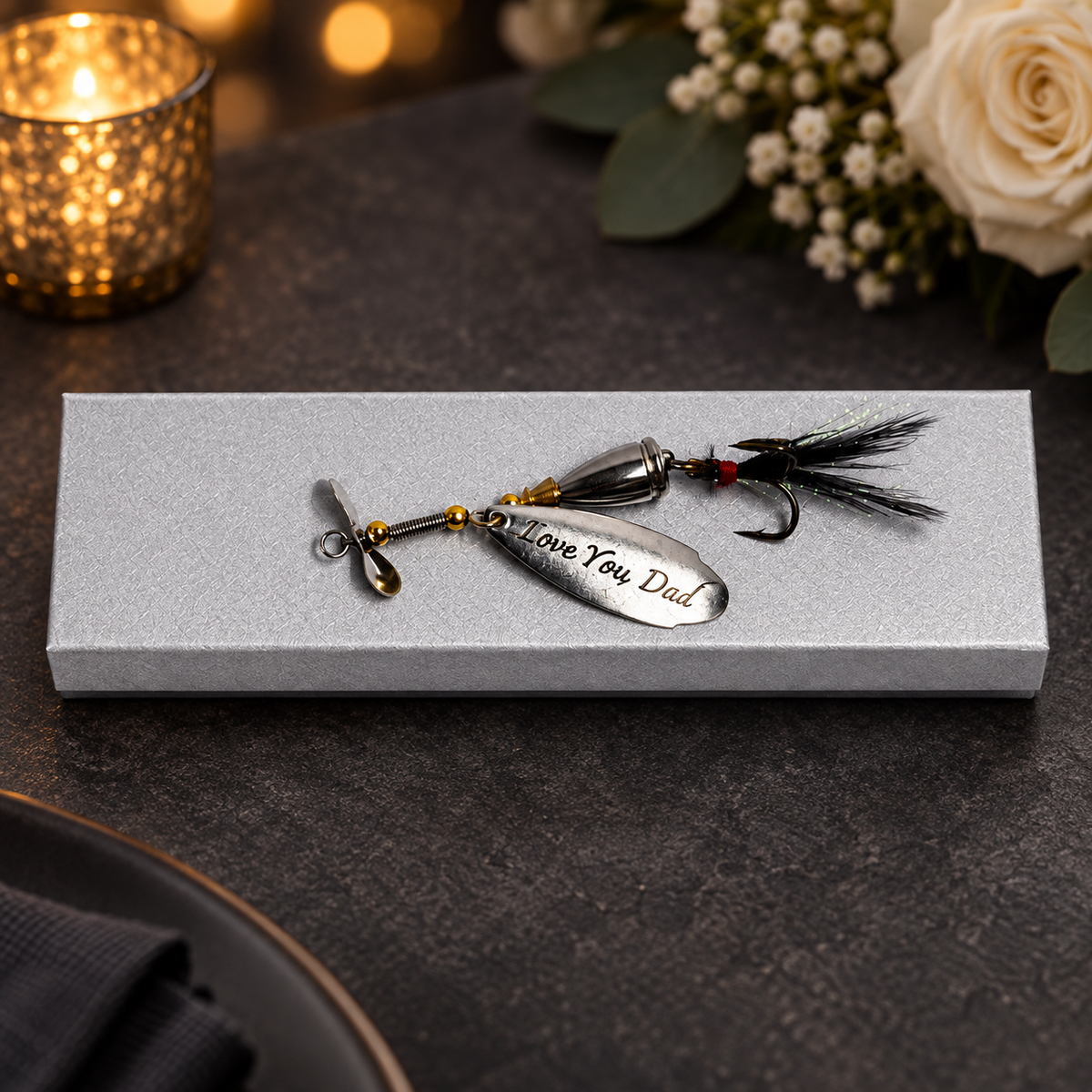 A silver metal spinnerbait fishing lure engraved with 'I Love You Dad' rests on a textured gray gift box. The lure features a black and red feathered tail and is photographed in a warm, styled setting with a glowing candle and flowers in the background.