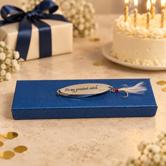A silver metal fishing lure engraved with 'To my greatest catch' rests on a textured blue gift box. The scene is styled for a birthday or celebration, with a cream-colored cake with lit candles, a wrapped gift box with a navy ribbon, and gold confetti scattered on a beige stone surface.